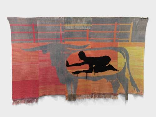 Zu sehen ist ein gewebter Teppich, der in Orange- und Rottönen gehalten ist. In der Mitte ist eine graue Stiersilhouette zu sehen, in dessen Mitte sich die Silhouette eines knieenden und gebückten Menschen eingerahmt ist.